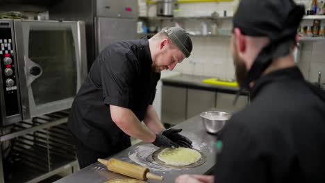 Ein-Selbstbewusster-Männlicher-Koch-In-Schwarzer-Uniform-Erklärt-Einem-Angehenden-Kollegen,-Wie-Man-Die-Soße-Richtig-Auf-Dem-Pizzateig-Verteilt,-Um-In-Der-Restaurantküche-Eine-Gute-Pizza-Vom-Pizzabäcker-Zuzubereiten.-Ein-Selbstbewusster-Männlicher-Pizzabäcker-In-Schwarzer-Uniform-Wirft-Pizza-Zum-Backen-In-Den-Ofen.