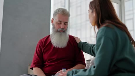 An-adult-brunette-girl-in-a-green-jacket-talks-with-her-dad-an-elderly-man-with-gray-hair-and-a-lush-beard-in-a-red-T-shirt-bout-his-health-in-a-modern-apartment