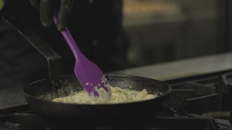 Close-up-a-professional-chef-in-a-black-uniform-stirs-the-contents-of-a-hot-frying-pan-and-adds-pieces-of-fish-while-cooking-a-dish-on-a-hotplate-in-a-restaurant