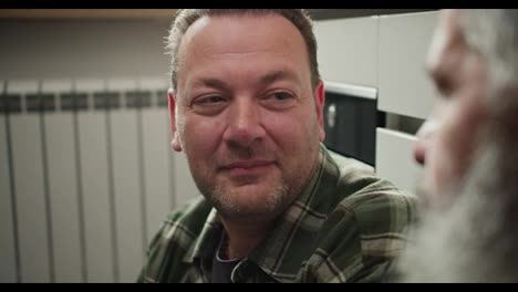 And-close-up-a-brunette-man-with-Universe-stubble-in-a-checkered-shirt-communicates-and-looks-at-his-elderly-boyfriend-with-a-gray-beard-sitting-on-the-floor-in-the-kitchen.-An-elderly-man-strokes-the-cheek-of-his-boyfriend-with-stubble-in-an-LGBT-couple