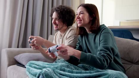 Chica-Morena-Feliz-Junto-Con-Su-Novio-Con-Cabello-Rizado-Envuelto-En-Una-Manta-Y-Jugando-Videojuegos-Mientras-Están-Sentados-En-El-Sofá-Usando-Un-Joystick-En-Casa