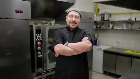 Portrait-of-a-confident-professional-male-chef-in-a-hat-and-black-uniform-with-a-beard-who-folds-his-arms-on-his-chest-and-feels-confident-in-a-restaurant-kitchen