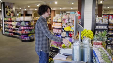 Happy-guy-with-curly-hair-in-a-plaid-shirt-weighs-bananas-using-scales-in-a-supermarket-while-shopping-in-a-grocery-store