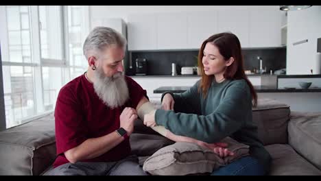 An-elderly-man-with-gray-hair-and-a-lush-beard-in-a-red-T-shirt-helps-his-adult-brunette-daughter-in-a-Green-jacket-bandage-his-hand-with-bandages-while-sitting-on-the-sofa-in-a-modern-apartment