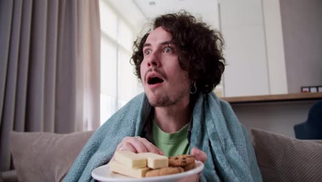 A-surprised-and-happy-brunette-guy-with-curly-hair-is-wrapped-in-a-blue-blanket-and-eats-sweet-snacks-while-watching-an-interesting-movie-at-home-on-the-couch