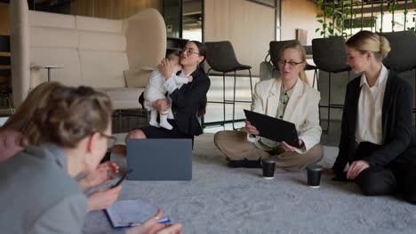 Close-up-five-confident-business-women-in-business-suits-sit-on-the-carpet-in-a-modern-office-and-communicate-while-one-of-them-holds-a-small-child-in-her-arms