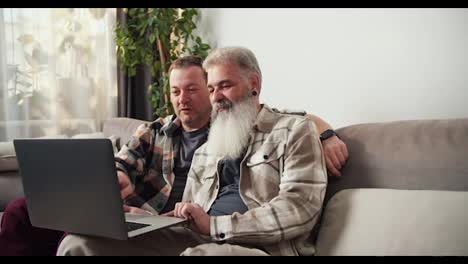 A-happy-middle-aged-man-with-gray-hair-and-a-lush-beard-sits-on-the-sofa-with-his-brunette-boyfriend-and-looks-at-the-monitor-of-a-gray-laptop-in-a-modern-apartment