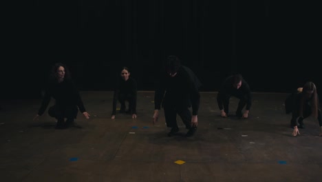 Five-confident-theatergoers-actors-stand-in-a-row-in-black-theater-clothes-and-practice-synchronized-movement-during-their-preparation-and-rehearsal-in-the-theater-on-stage-near-black-curtains