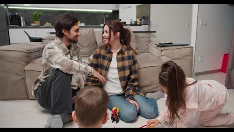 Un-Hombre-Moreno-Con-Barba-Incipiente-Y-Camiseta-Beige-Se-Comunica-Con-Su-Esposa,-Que-Lleva-Una-Camisa-A-Cuadros,-Sentada-En-El-Suelo-Mientras-Sus-Hijos-Hacen-Creatividad-Y-Dibujan-En-Hojas-De-Papel-Sentados-En-El-Suelo-Y-Se-Divierten-En-Un-Nuevo-Y-Moderno-Apartamento.