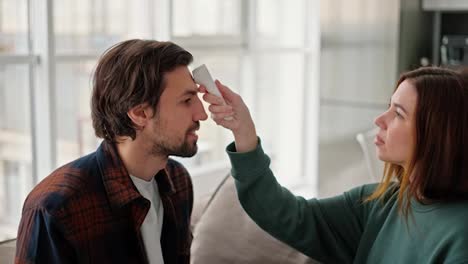 Ein-Glückliches-Brünettes-Mädchen-In-Einem-Grünen-Pullover-Misst-Die-Temperatur-Ihres-Brünetten-Freundes-Mit-Stoppeln-In-Einem-Orange-blauen-Hemd-Auf-Dem-Sofa-In-Einem-Modernen-Apartment