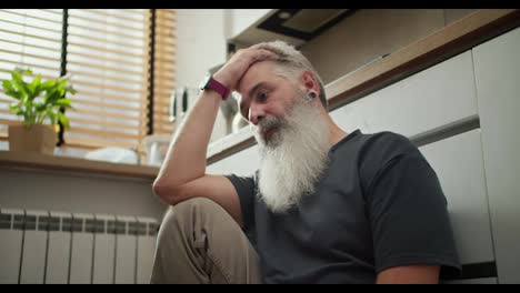 A-serious-man-with-gray-hair-and-a-lush-gray-beard-in-a-gray-T-shirt-sits-on-the-floor-and-holds-his-head-with-one-leg-in-the-kitchen.-A-thoughtful-and-serious-elderly-man-sits-in-the-kitchen-and-looks-at-the-floor-while-holding-his-forehead-with-his-hand