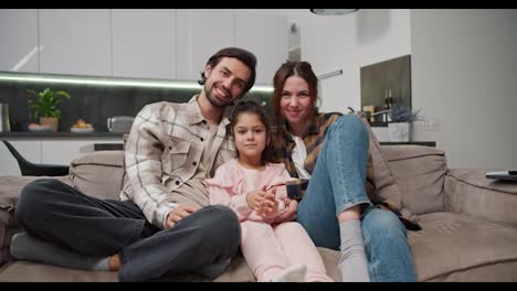 Portrait-of-a-happy-family,-a-brunette-man-with-stubble-along-with-his-brunette-wife-in-a-checkered-shirt-sitting-on-a-brown-sofa-along-with-their-little-daughter-in-a-pink-suit-in-a-modern-new-apartment