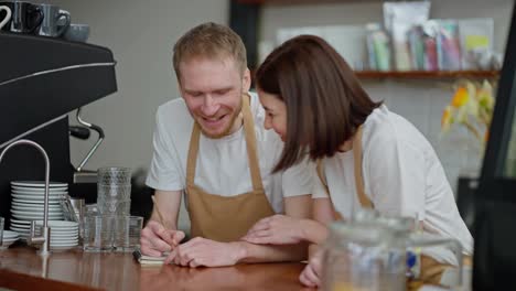 Happy-blond-guy-in-light-brown-apron-with-his-colleague-waitress-brunette-girl-making-up-their-plan-and-writing-down-the-main-ideas-while-working-at-the-counter-in-a-cafe