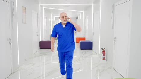 A-confident-doctor-with-a-gray-beard-in-glasses-and-a-blue-uniform-hurries-and-walks-along-the-corridor-of-a-modern-clinic-communicating-on-a-white-smartphone