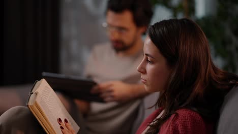 Side-view-of-a-confident-brunette-girl-in-a-red-sweater-reading-a-book-while-sitting-on-the-sofa-with-her-boyfriend-who-is-busy-with-a-tablet-in-a-modern-apartment