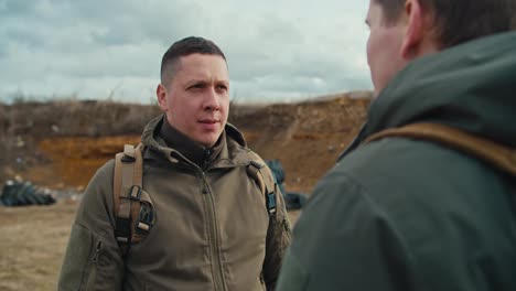 Over-his-shoulder-a-confident-military-man-communicates-with-his-colleague-in-a-dark-green-army-uniform-and-a-backpack-on-his-shoulders-on-the-territory-of-a-military-training-ground