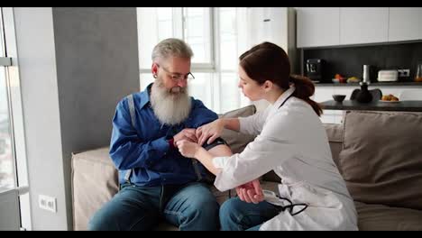 A-brunette-woman-doctor-in-a-white-coat-installs-a-special-instrument-on-a-mans-shoulder-to-measure-pulse-and-pressure-during-an-examination-at-home-of-an-elderly-man-with-gray-hair-and-a-lush-beard-in-a-blue-shirt-on-a-sofa-in-a-modern-apartment