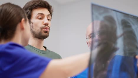 A-couple-of-confident-doctors-a-girl-and-an-elderly-man-with-a-gray-beard-and-glasses-reassure-a-guy-showing-good-results-of-fluorography-on-an-image-in-a-modern-clinic