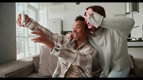 Happy-young-man-with-Black-skin-color-in-a-checkered-cream-shirt-takes-a-selfie-with-his-brunette-girlfriend-in-a-sweater-using-a-white-smartphone-while-sitting-on-a-modern-sofa-in-a-studio-apartment