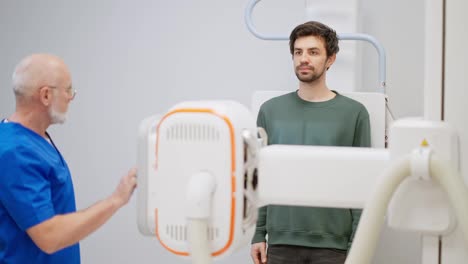 Confident-brunette-guy-with-stubble-in-a-green-jacket-undergoing-fluorography-by-an-elderly-experienced-doctor-in-a-blue-uniform-in-a-modern-bright-clinic