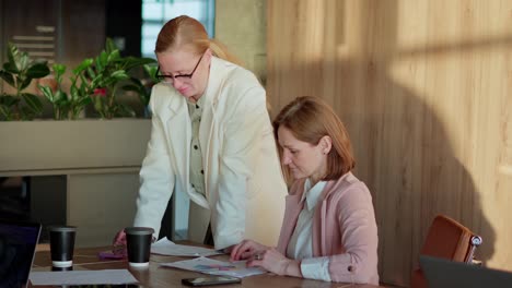 A-confident-middle-aged-blonde-girl-in-a-white-suit-together-with-her-businesswoman-colleague-gathered-at-the-table-in-the-office-and-solve-current-problems-and-plan-their-work