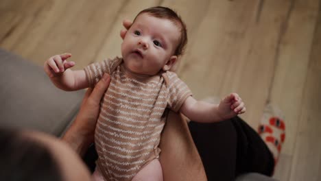 Close-up-from-above-a-happy-father-holds-his-little-baby-daughter-in-a-brown-overalls-in-his-arms-in-a-modern-apartment-at-home