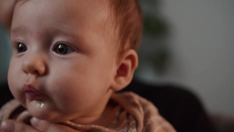 Primer-Plano-De-Una-Pequeña-Niña-Con-Ojos-Grandes-Mirando-A-La-Cámara-Y-Posando-En-Un-Apartamento-Moderno-Sentada-En-Los-Brazos-De-Su-Madre.