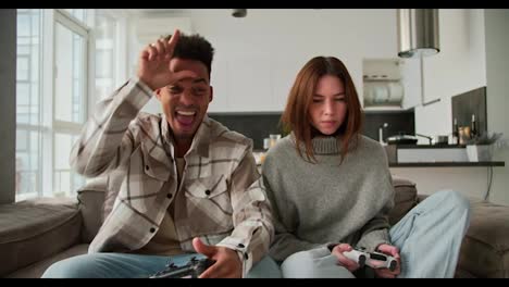 Happy-and-cheerful-man-with-Black-skin-color-a-young-brunette-with-stubble-in-a-cream-checkered-shirt-is-very-happy-about-his-Victory-in-a-video-game-while-sitting-with-his-girlfriend-in-a-sweater-on-a-gray-sofa-in-a-modern-apartment