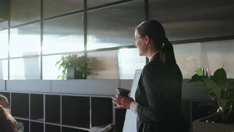 Brunette-businesswoman-in-business-clothes-holds-a-glass-of-coffee-in-her-hands-and-tells-her-colleagues-about-her-ideas-using-a-stand-in-a-sunny-office-during-a-meeting