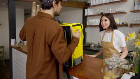 Happy-girl-waiter-in-a-yellow-apron-greets-a-man-delivering-food-and-gives-him-a-finished-order-in-a-cafe