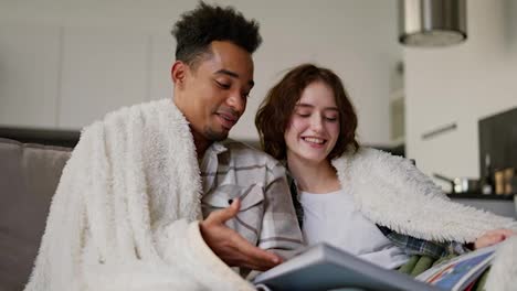 A-happy-young-man-with-Black-skin-color-a-brunette-in-a-checkered-beige-shirt-is-sitting-on-the-sofa-with-his-young-adult-brunette-girlfriend-with-a-bob-hairstyle-wrapped-in-a-white-woolen-blanket.-They-are-reading-a-book-and-relaxing-in-a-modern-apartment