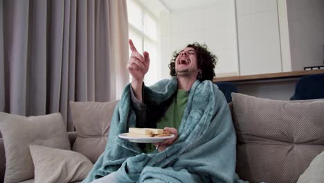 Happy-cheerful-brunette-guy-with-curly-hair-in-a-blue-plaid-actively-laughs-and-eats-sweet-snacks-while-watching-a-funny-movie-and-comedy-at-home-in-a-modern-apartment