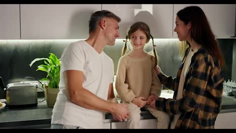 Happy-little-brown-haired-girl-sits-on-the-table-in-the-kitchen-and-communicates-with-her-brunette-mom-in-a-plaid-shirt-and-middle-aged-dad-with-gray-hair-in-a-white-T-shirt-in-a-modern-kitchen-in-the-evening