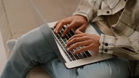 Close-up-of-a-young-man-with-Black-skin-color-in-a-beige-plaid-shirt-and-blue-jeans-works-on-a-gray-laptop-and-types-on-the-keyboard-while-working-remotely-at-home-on-the-sofa