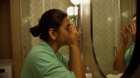 Side-view-of-a-happy-brunette-girl-in-green-pajamas-making-an-evening-mask-and-applying-special-oils-to-her-face-in-a-modern-apartment-before-going-to-bed-in-the-evening-in-the-bathroom