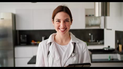 Portrait-of-a-happy-brunette-girl-in-a-white-doctors-coat-with-a-stethoscope-on-her-shoulders-and-with-a-tablet-in-her-hands-near-a-modern-kitchen-in-an-apartment-for-a-home-examination