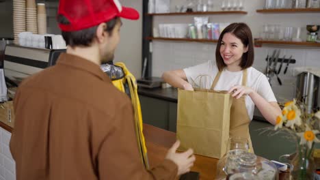 Eine-Brünette-Barista-Gibt-Einem-Mann,-Der-Essen-In-Einem-Café-Ausliefert,-Eine-Fertige-Bestellung.-Ein-Mann,-Der-Essen-Ausliefert,-Kam,-Um-Eine-Bestellung-In-Einem-Café-Abzuholen