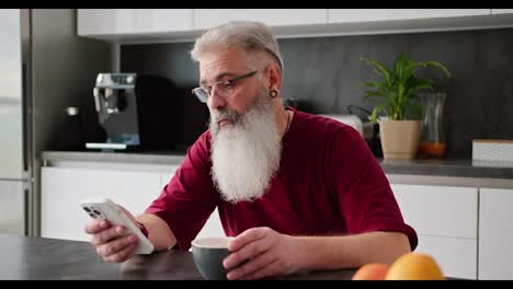 Un-Hombre-Serio,-De-Pelo-Gris-Y-Barba-Frondosa,-De-Edad-Avanzada,-Con-Gafas-Y-Camisa-Roja,-Se-Sienta-A-Mirar-Una-Transmisión-En-Un-Teléfono-Inteligente-De-Una-Red-Social-Durante-Su-Desayuno-Y-Sonríe-En-Una-Cocina-Moderna.