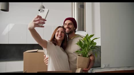 Happy-brunette-girl-in-a-white-t-shirt-hugging-her-brunette-boyfriend-in-a-red-hat-and-cream-t-shirt-who-is-holding-a-house-plant-and-taking-a-selfie-with-her-girlfriend-in-her-new-modern-apartment-after-moving-with-boxes-around