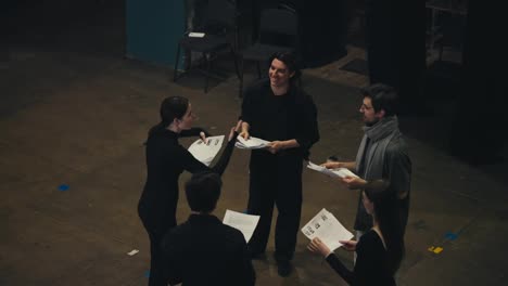 Top-view-of-a-group-of-confident-actors-in-black-suits-Standing-in-a-circle-and-talking-to-each-other-holding-sheets-of-paper-with-a-script-in-their-hands-and-talking-together-with-their-director-about-the-play-and-performance-on-stage-in-the-theater