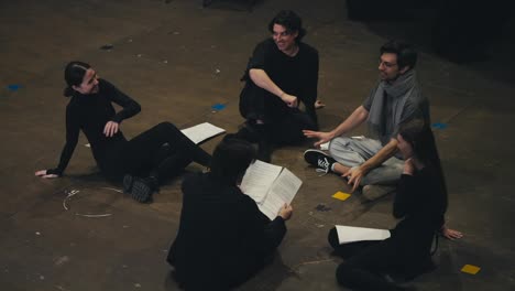 A-group-of-confident-young-actors-in-black-suits-are-sitting-on-stage-together-with-their-director-holding-sheets-of-paper-in-their-hands-with-the-script-of-the-play-on-stage-in-the-theater