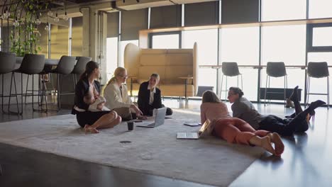 Side-view-of-a-group-of-businesswoman-in-business-clothes-lying-on-the-carpet-in-a-modern-office-talking-and-during-a-break-between-work-a-brunette-girl-breastfeeds-her-little-child-in-the-office