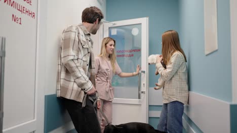 Una-Veterinaria-Con-Uniforme-Rosa-Invita-A-Una-Chica-Rubia-Con-Un-Perro-Blanco-A-Entrar-A-La-Oficina-De-La-Clínica-Veterinaria-Mientras-El-Chico-Con-Su-Perro-Negro-Se-Queda-En-El-Pasillo-Esperando-Su-Turno.