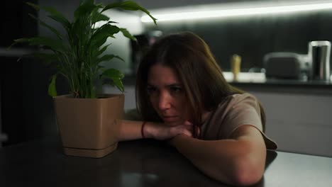 A-sad-and-thoughtful-brunette-girl-in-a-beige-T-shirt-sits-leaning-on-a-black-dining-table-in-the-kitchen-near-a-green-indoor-plant-in-a-pot-in-the-evening-in-a-modern-kitchen