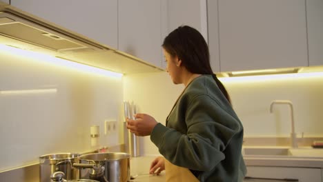 Confident-brunette-girl-in-a-green-jacket-and-beige-apron-prepares-soup-and-adds-the-necessary-spice-ingredients-to-the-pan-while-cooking-at-home-in-a-modern-apartment