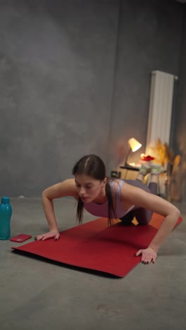 Vertical-video-Confident-brunette-girl-in-sports-summer-uniform-doing-push-ups-on-a-red-sports-mat-near-a-sports-bottle-of-water-in-a-modern-apartment-at-home