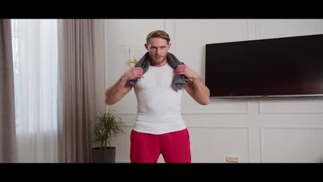 Portrait-of-a-confident-athletic-blond-man-with-a-beard-in-a-white-T-shirt-with-a-gray-towel-around-his-neck-after-his-exercise-and-workout-at-home-in-a-modern-apartment