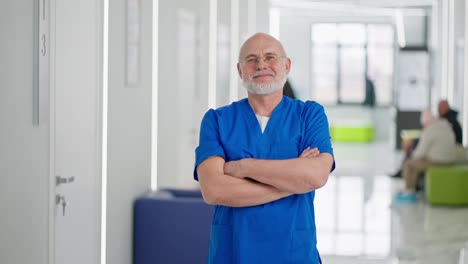 Retrato-De-Un-Médico-Mayor,-Confiado-Y-Feliz,-Con-Gafas,-Barba-Gris-Y-Uniforme-Azul,-Que-Cruzó-Los-Brazos-Sobre-El-Pecho-Y-Posó-Con-Confianza-En-El-Pasillo-De-Una-Clínica-Moderna.