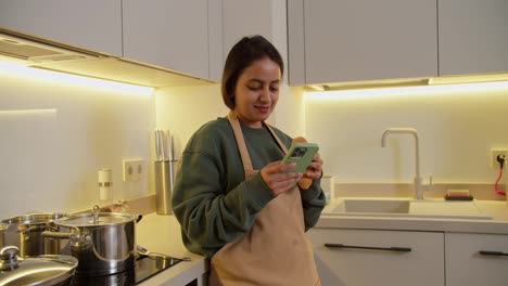 Happy-brunette-Indian-girl-in-a-Green-sweater-and-beige-apron-eats-a-croissant-and-types-on-her-green-smartphone-on-the-social-network-during-her-break-and-lunch-at-home-in-a-modern-apartment