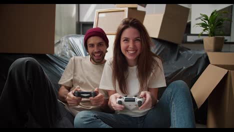 Una-Chica-Morena-Feliz-Con-Una-Camiseta-Blanca-Juega-Videojuegos-Con-Su-Novio-Usando-Joysticks-Y-Se-Divierte-Mucho-Sentada-En-El-Suelo-Cerca-Del-Sofá,-Envuelto-En-Plástico-Negro-Y-Con-Muchas-Cajas-Alrededor-Después-De-Mudarse.